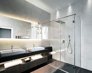Modern bathroom with LED strip lighting above mirror, shower with spotlights, light gray wall tiles, dark gray floor tiles, black vanity unit with twin sinks