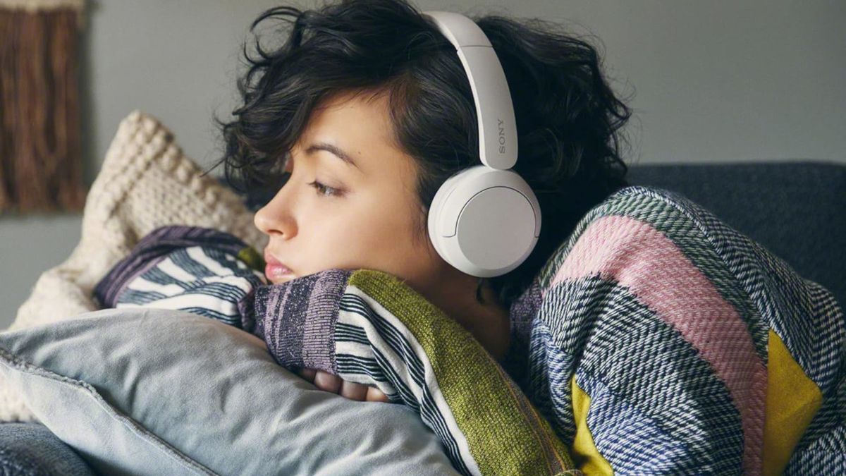 Best budget wireless headphones: Woman with black hair wearing a colourful shirt, resting on cushion on her sofa and wearing a pair white Sony headphones