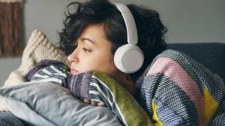 Woman with black hair wearing a colourful shirt, resting on cushion on her sofa and wearing a pair white Sony headphones