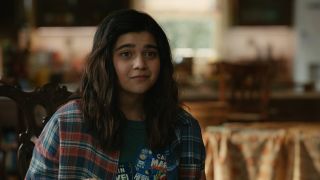 Still from The Marvels (2023) movie. Close up of Kamala Khan sitting on a dining chair at her home, with the kitchen in the background. She is a teenager with long dark hair and is wearing a Captain Marvel-motif t-shirt and blue and red plaid shirt over top. She has a slight smirk on her face.