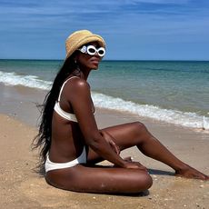 Woman on beach wears white swimsuit, white sunglasses and raffia hat