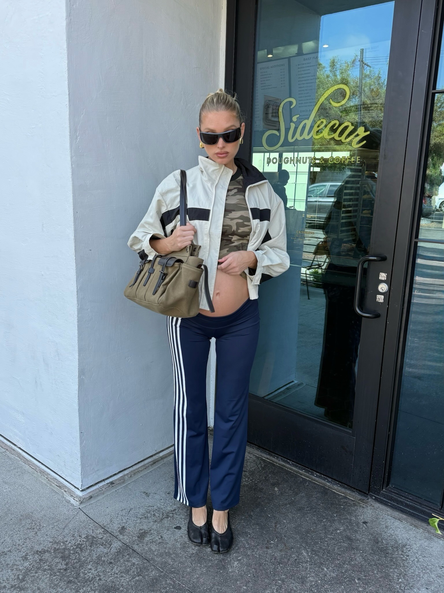 Elsa Hosk wearing a windbreaker, a camo T-shirt, Adidas track pants, Maison Margiela Tabi ballet flats, and a Prada Route bag.