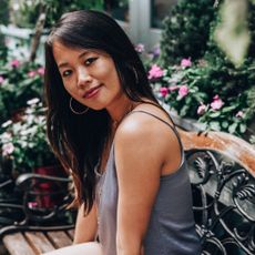 A portrait of the author Ana Huang, sitting on a wooden bench in a garden.