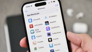 Photograph of App Shortcuts open on an iPhone 14 Pro with an iPad and AirPods visible in the background.
