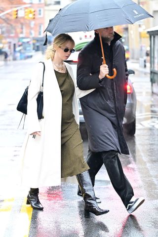 Jennifer lawrence wears a long white coat, black shoulder bag, green checkered maxi dress, and black croc heeled boots.