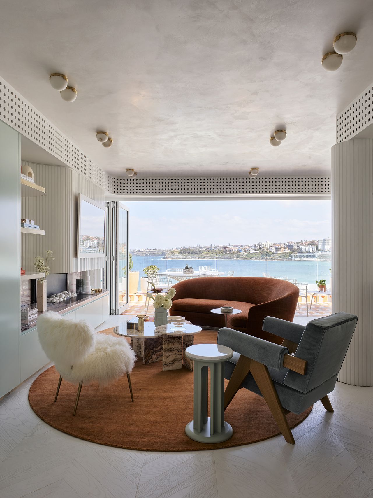 70s style living room with rust orange sofa, orange rug, mint green cabinetry, fluted surface above marble fireplace with mirror/tv, shaggy armchair, blue velvet armchair, perforated detail along the roofline concealing aircon