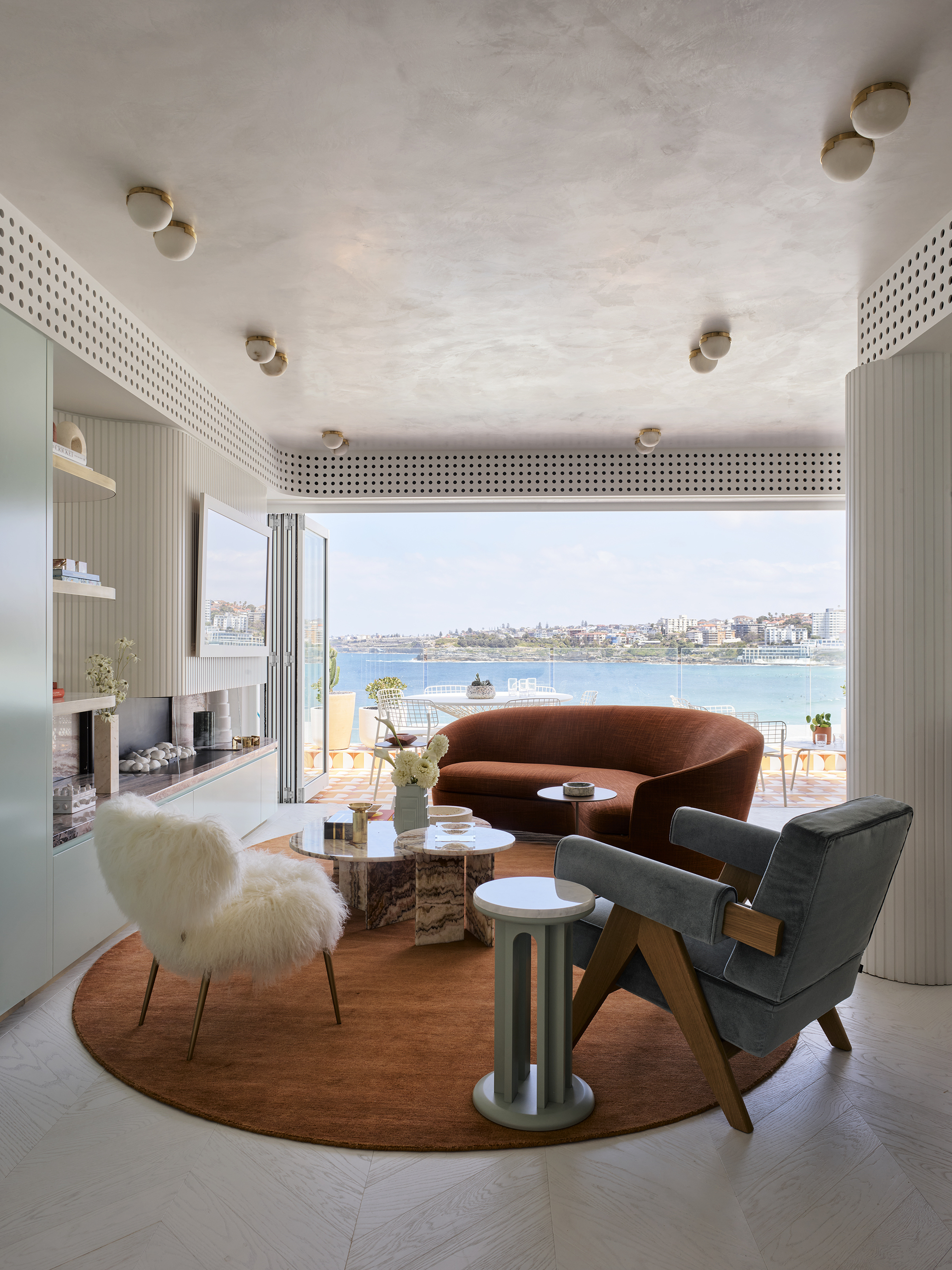 70s style living room with rust orange sofa, orange rug, mint green cabinetry, fluted surface above marble fireplace with mirror/tv, shaggy armchair, blue velvet armchair, perforated detail along the roofline concealing aircon