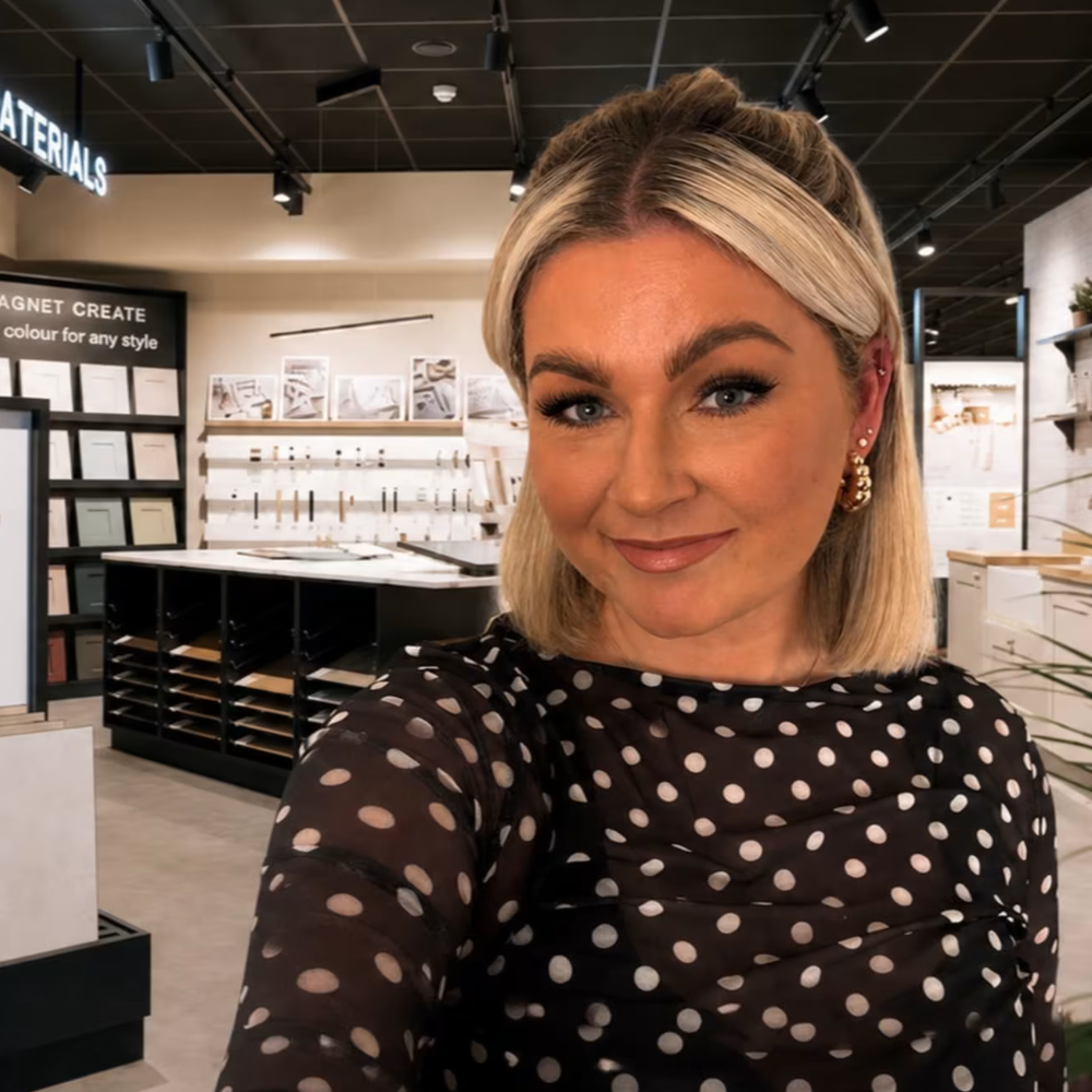 Smiling woman with blonde bob in black polka-dot top takes selfie in Magnet kitchen showroom with flooring samples