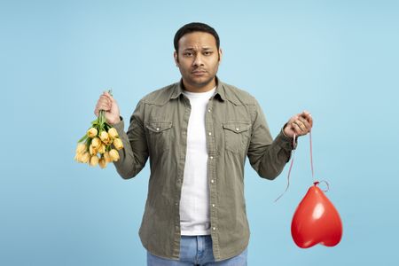 A sad man holding a deflated balloon and upside-down bouquet of flowers