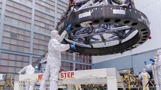 scientists in white clean suits move a metallic honeycomb-like structure onto a rectangular piece of electronics covered in wires and components