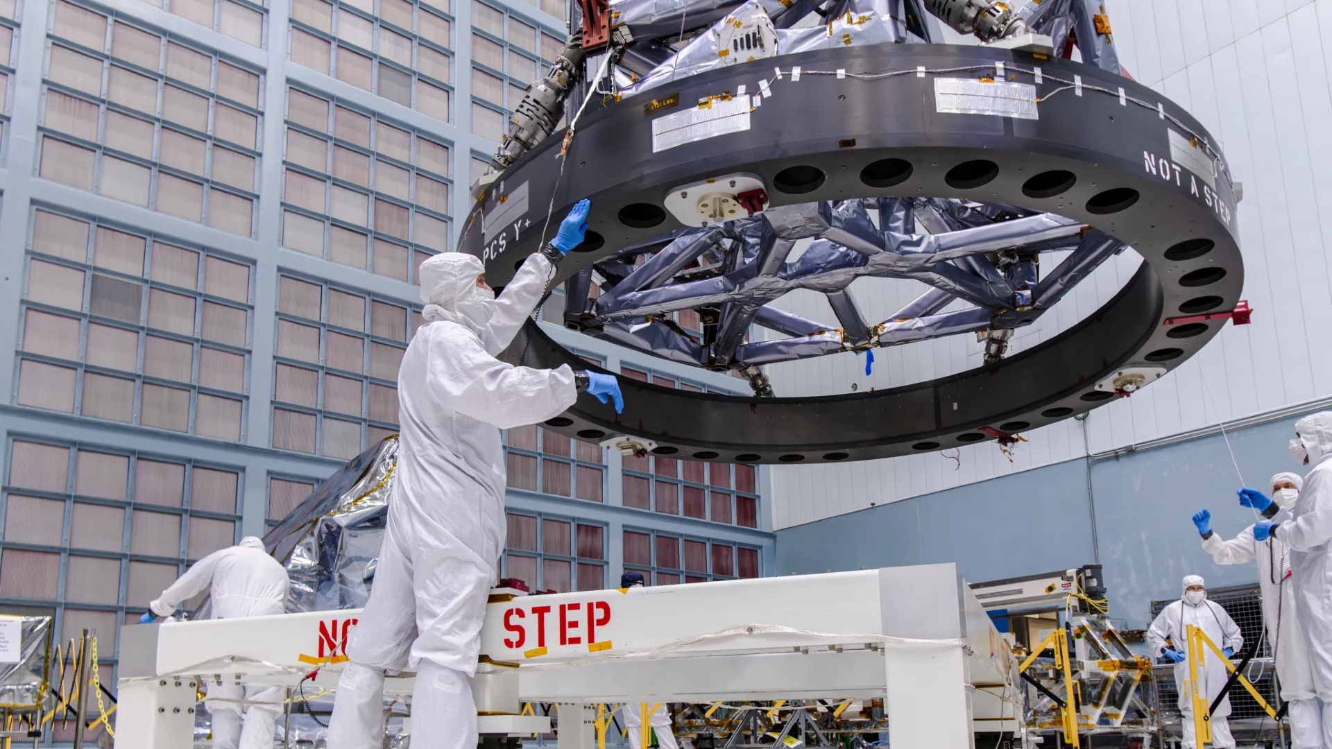 NASA Space Technology scientists in white clean suits move a metallic honeycomb-like structure onto a rectangular piece of electronics covered in wires and components