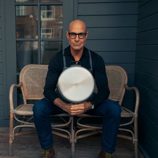 Stanley Tucci sitting on a bench outside of a house holding his stainless steel TUCCI by GreenPan pan