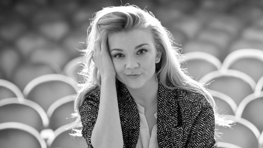 Black and white image of Natalie sitting in an empty theatre on stage, looking into the camera with a slight smile on her face 