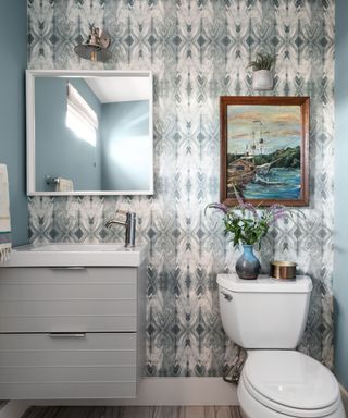 A blue bathroom with wallpaper, wall art, a mirror, a sink, and a toilet