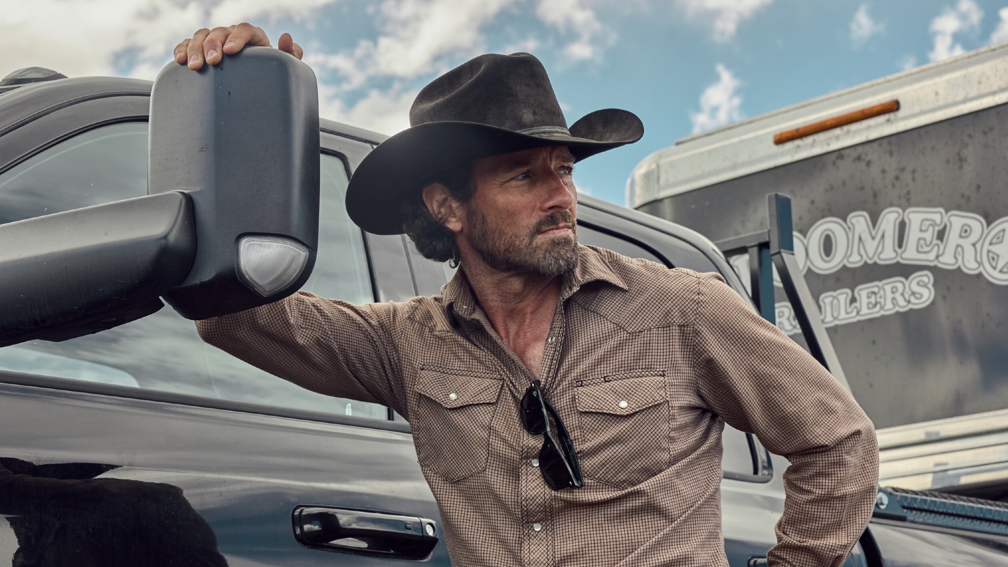Ian Bohen as Ryan resting his left hand on the mirror of a truck in Yellowstone.