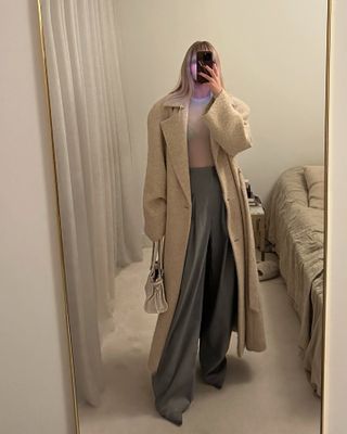 A woman takes a mirror selfie wearing a beige wool overcoat with gray trousers.