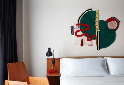  Ace Brooklyn hotel room, white walls, dark grey curtain, wooden sitting chair next to a white double bed, with wooden frame and side shelf, black spot lamp, colourful abstract artwork on wall over bed 