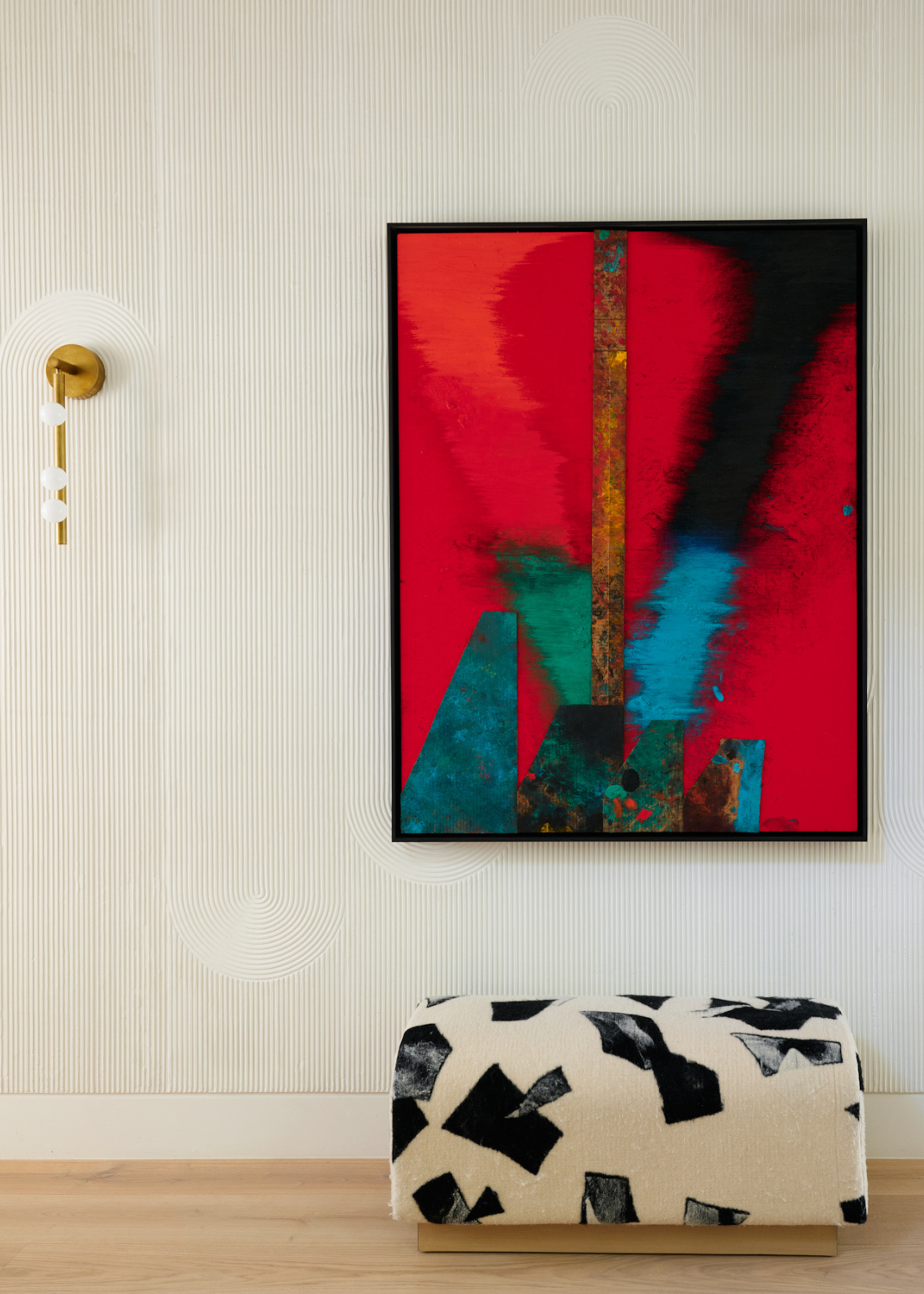 A living room with a framed colorful painting, a gold wall sconce with exposed bulbs, and a patterned bench