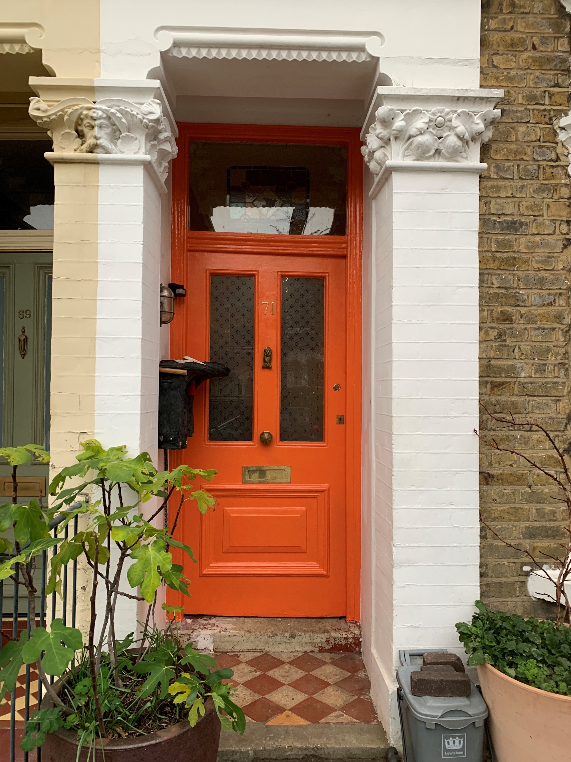 Image of a home with a bright orange front door. There is a red and cream tile front stoop, a black letterbox, and white trim around the alcove.
