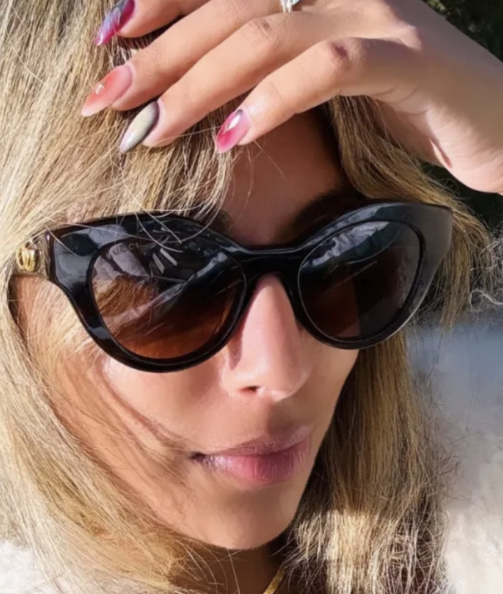 a white woman with dark sunglasses, blonde hair, and pink nails