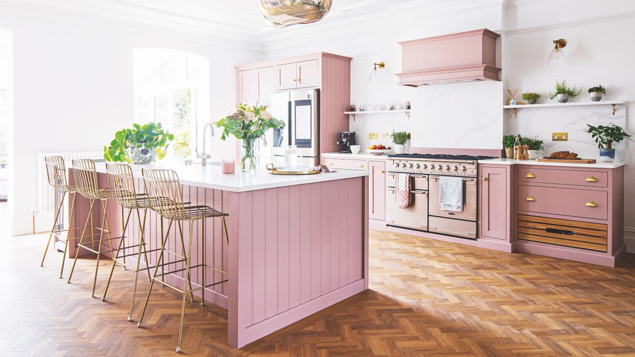 white kitchen with pink painted units, herringbone floor, brass bar stools, silver range cooker, vases of flowers, plants, open shelving 