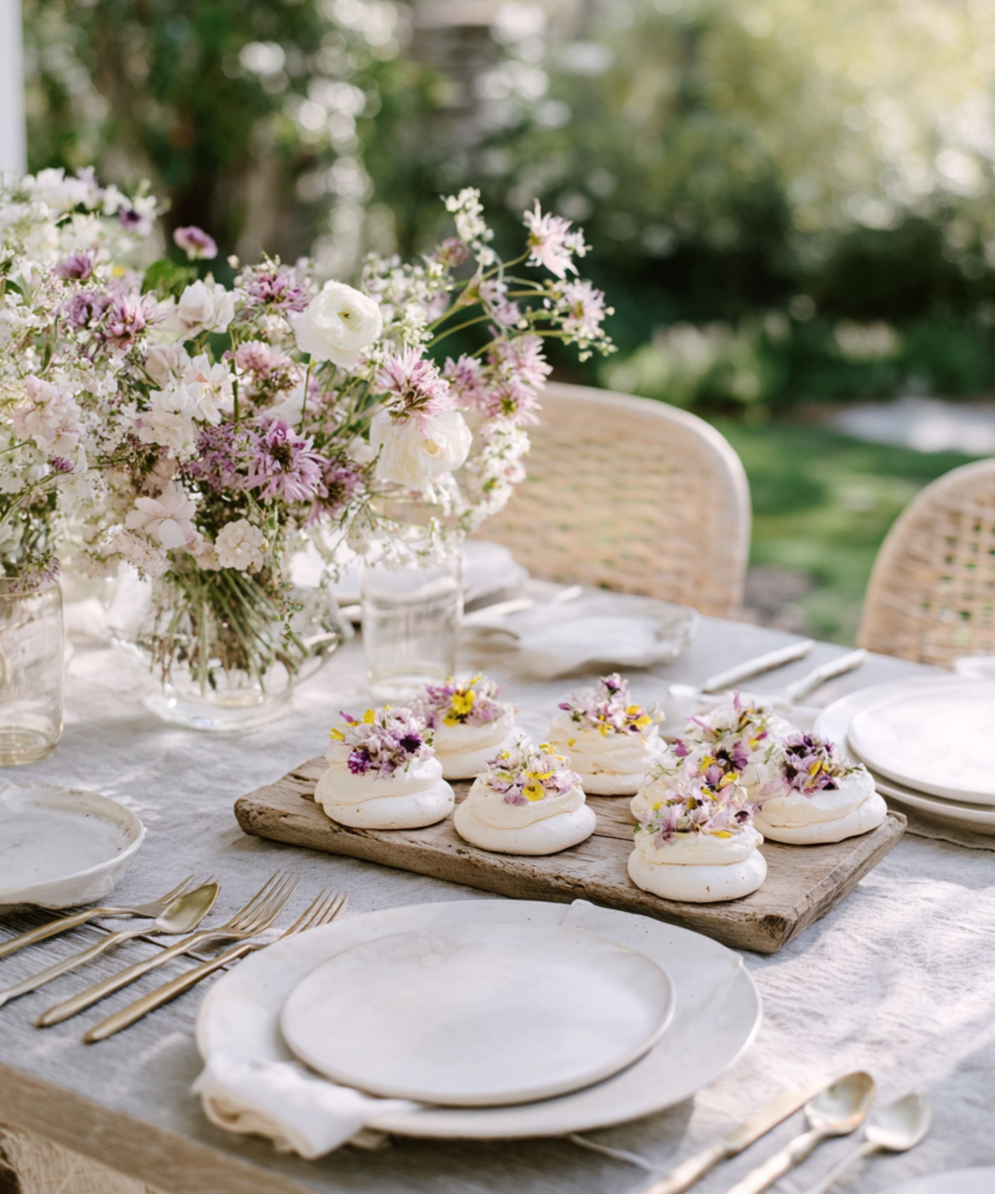 Mini meringues with edible flowers on a rustic outdoor dining table with linen tablecloth, rattan chairs and vases of flowers