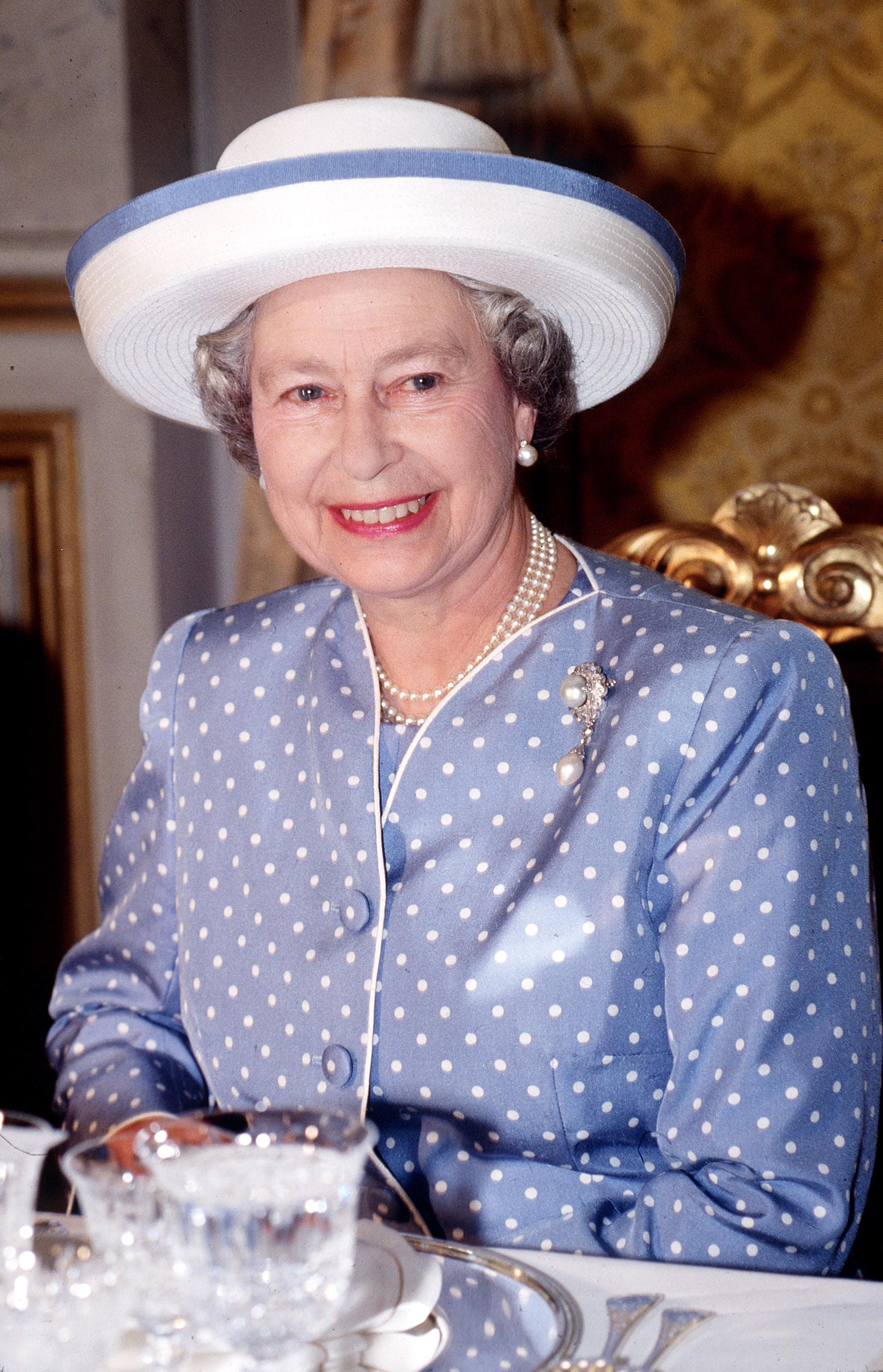 Queen Elizabeth wearing a lavender polka dot dress and white hate smiling at a dinner table