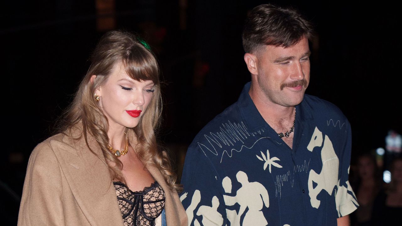 Taylor Swift wearing a tan coat and black bustier holding hands with Travis Kelce, wearing a blue patterned short sleeve shirt and smiling