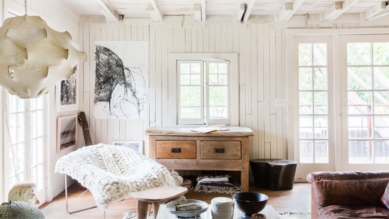 Living room with white shiplap walls, French doors, white beamed ceiling, wooden floor, a wooden console underneath the window, a metal armchair with a chunky knit throw on top and a leather sofa