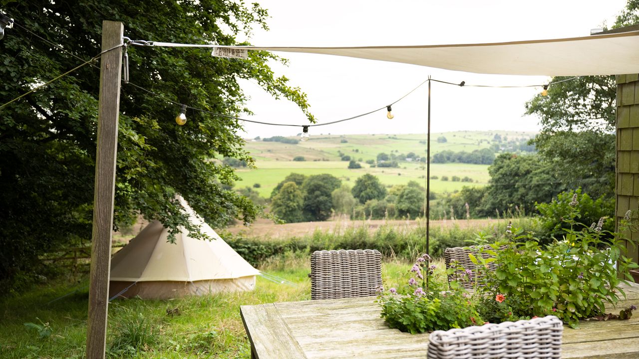 White canvas sail shade supported by a wooden post over a wooden garden dining table with wicker dining chairs