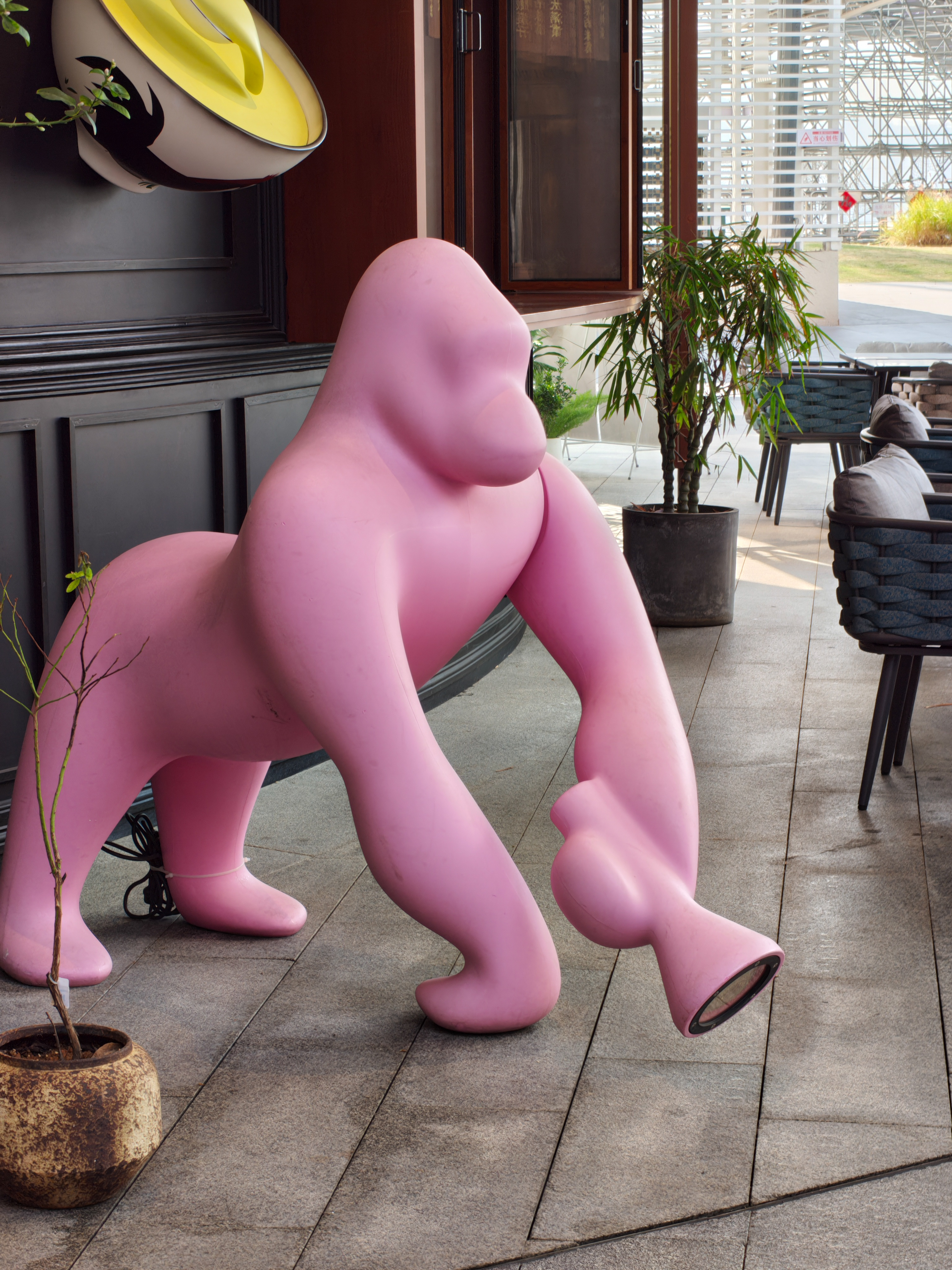 A large, smooth pink sculpture of a gorilla in a knuckle-walking pose. It is situated on a gray tiled outdoor walkway next to a dark-paneled building wall. In the background, a small potted bamboo plant and several modern patio chairs with gray cushions are visible near a white architectural structure.