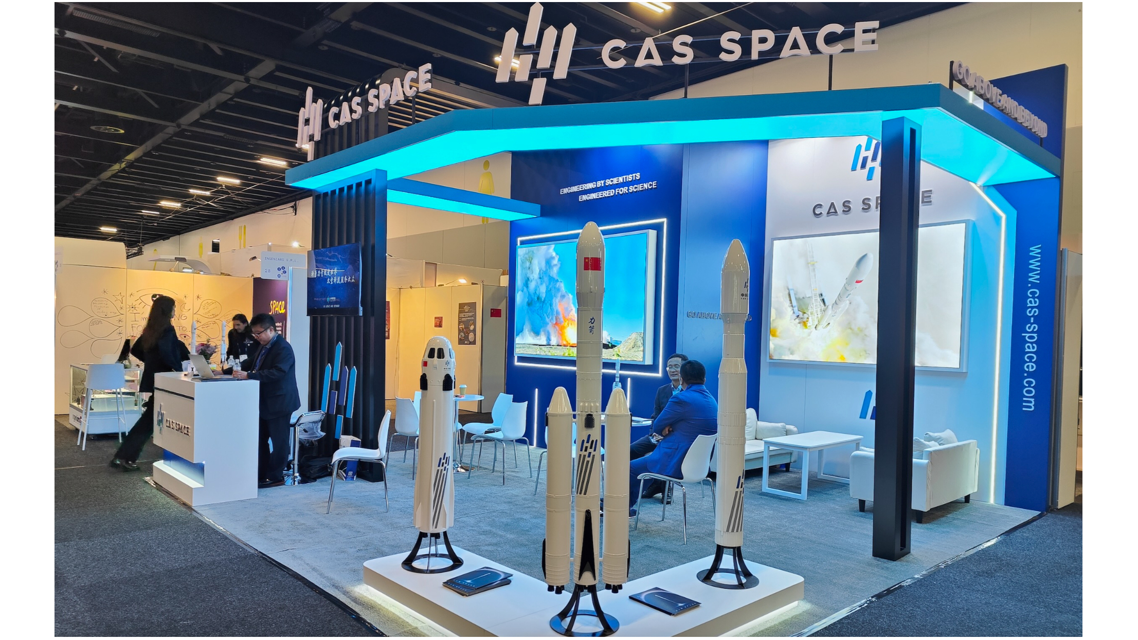 a booth in an exhibition hall at a space conference, showing three white model rockets on a white platform with a few people in the background