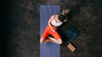 Woman seen from above sitting on yoga mat looking at smartphone with a cork yoga block next to her