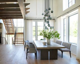 dining area beside staircase with statement pendant light, large wooden table, floor to ceiling windows, gray chairs and wooden floor