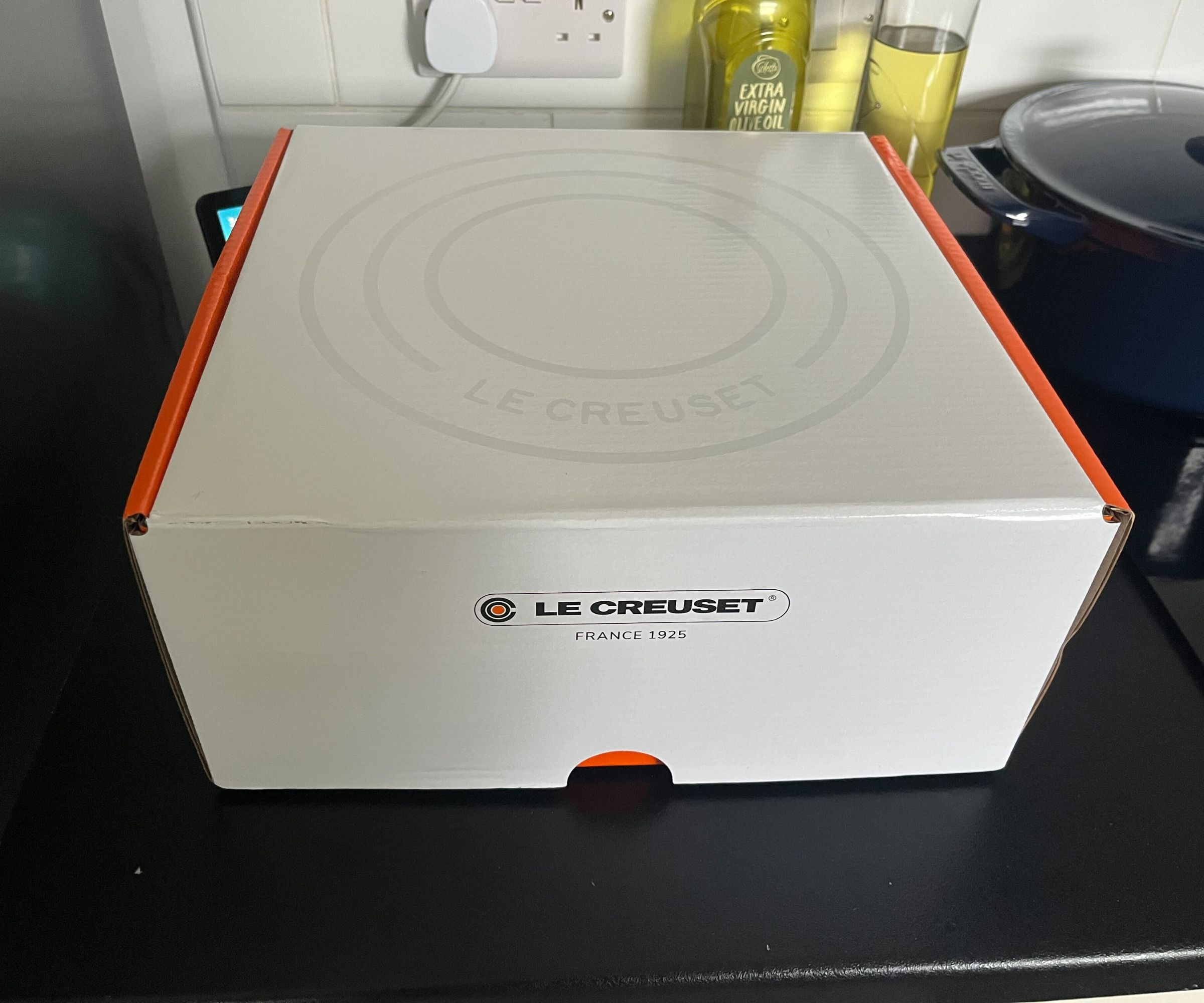 The white and orange Le Creuset Dutch oven box on a black kitchen counter with oil bottles in the background