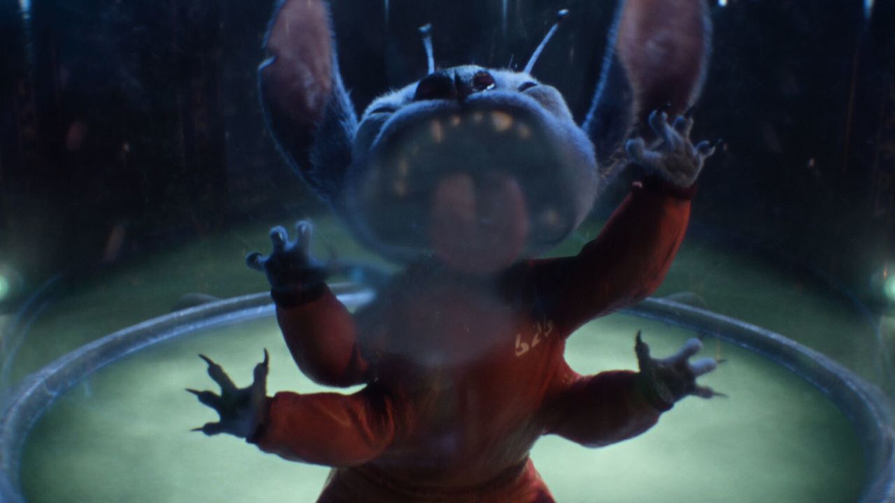 Still from the Lilo &amp; Stitch remake movie (2025). Stitch, a cute, furry blue alien with four arms and large ears, is wearing an orange prison jumpsuit. They are trapped inside a glass chamber. They are trying to claw their way out, licking the glass in the process.