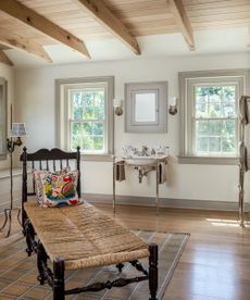 Bright neutral bathroom with wooden floors, ceiling beams and a wicker and mahogany chaise and white ceramic sink with steel stands and blue detailing