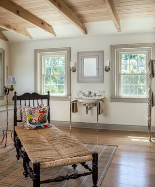 Bright neutral bathroom with wooden floors, ceiling beams and a wicker and mahogany chaise and white ceramic sink with steel stands and blue detailing