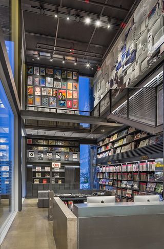 Interior view of the Hyundai Music Library featuring shelving and display units filled with records, a workspace with Apple Mac computers, DJ decks, an 'ON' sign and glass windows. The upper level can also be seen which features colourful wall art on one wall and another wall that is filled with painted electrical items