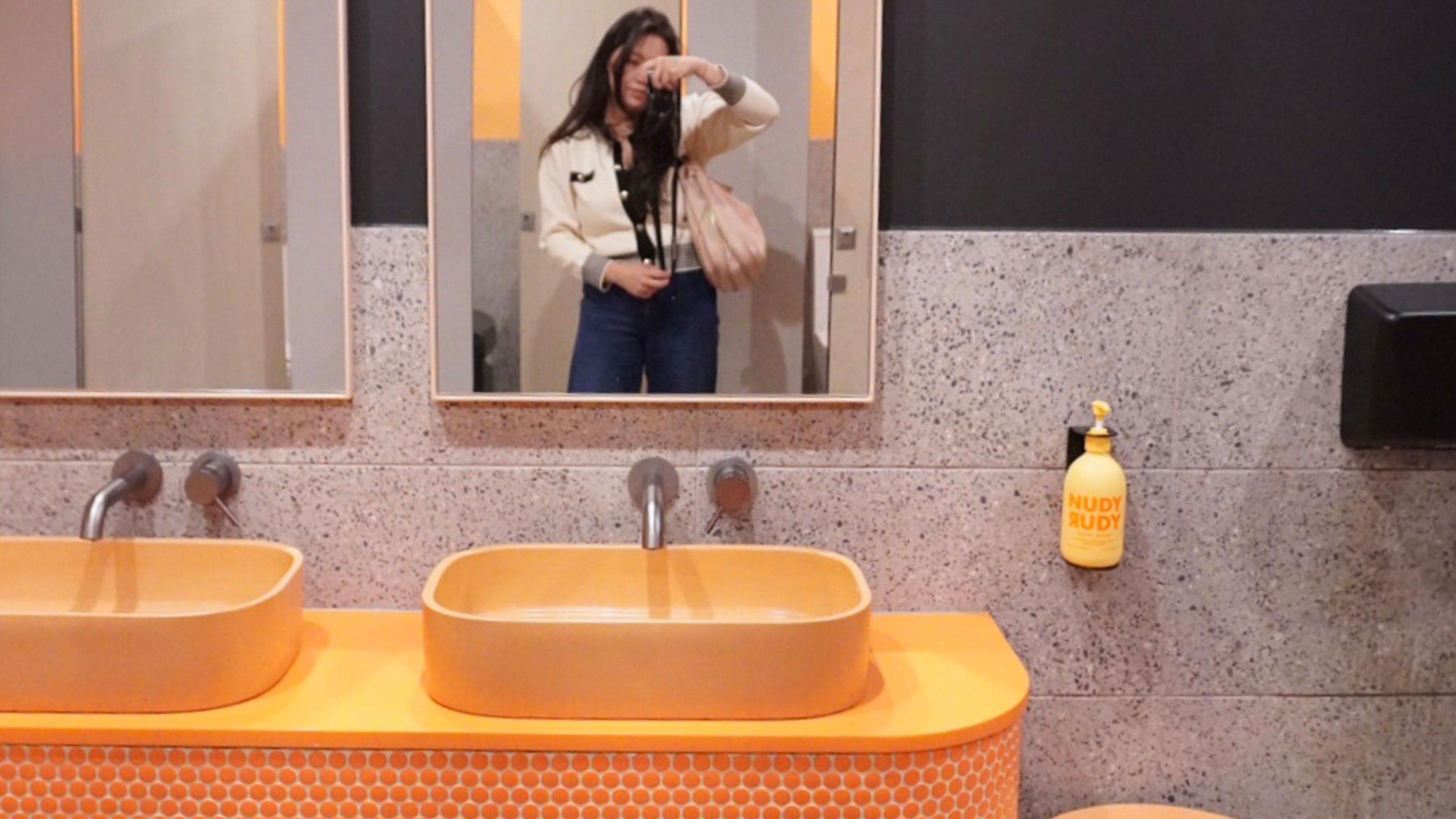 Woman taking selfie in bathroom mirror above orange sinks and countertops 