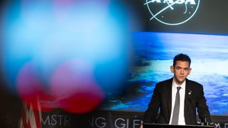 a man in a dark suit speaks at a lectern in front of an image of earth on a large screen behind him