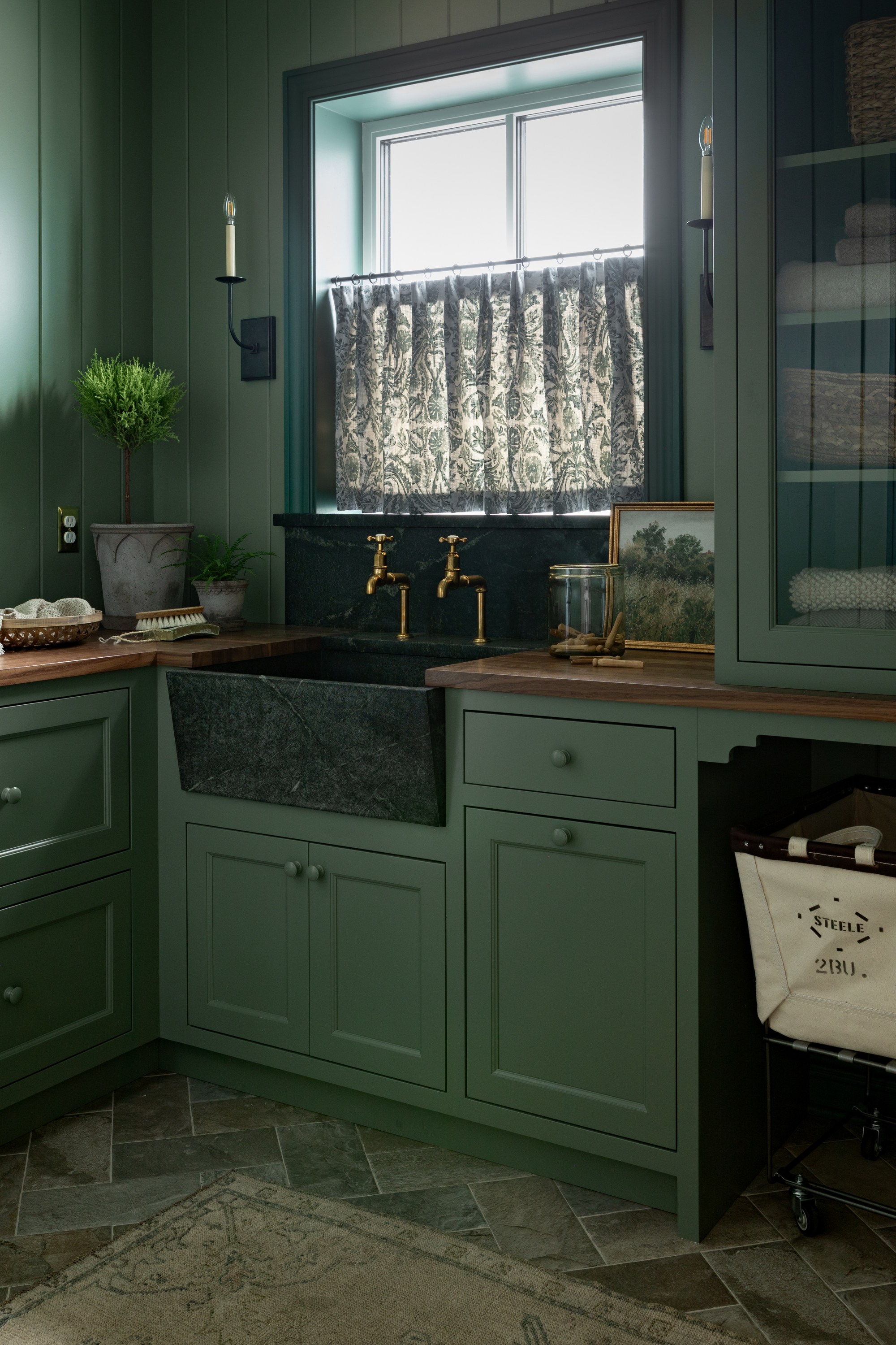 Image of dark laundry room color drenched in a mid-tone green paint color. There are wooden countertops and gold finishes. There is a cafe curtain on the window above the sink.