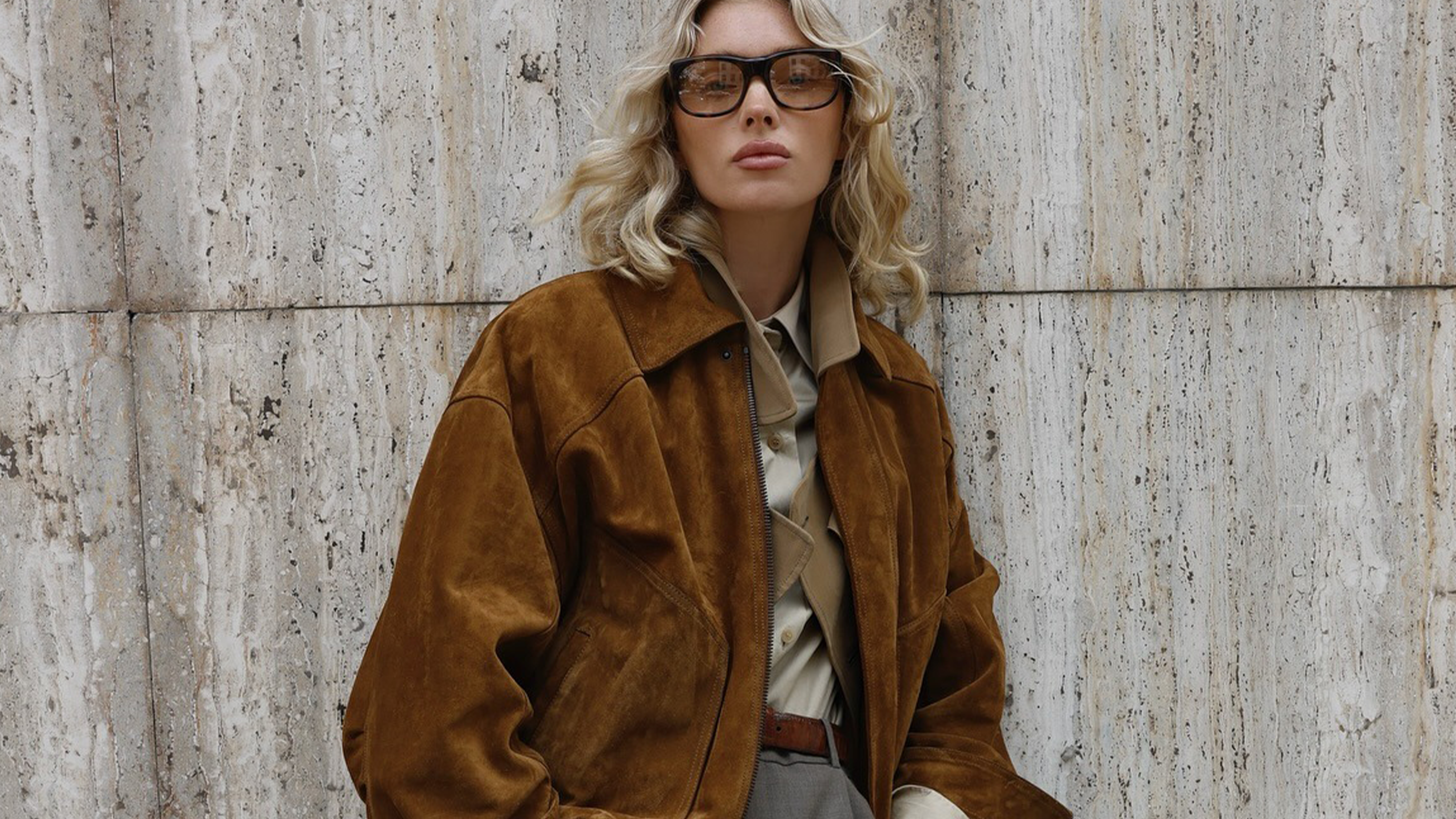 Model Elsa Hosk wearing suede jacket sunglasses and gray pants standing against stone background.