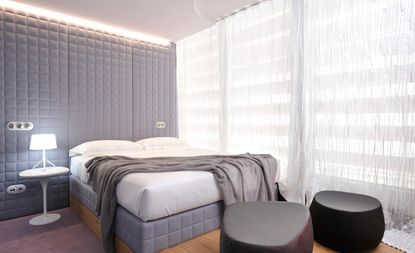 Hotel room, grey padded wall, double bed in front with grey padded base and white duvet cover and pillows, grey bed throw, wooden platform, purple carpet with white bedside table and lit lamp, white voile curtains inform of large windows, a black and a purple bean bag style seats