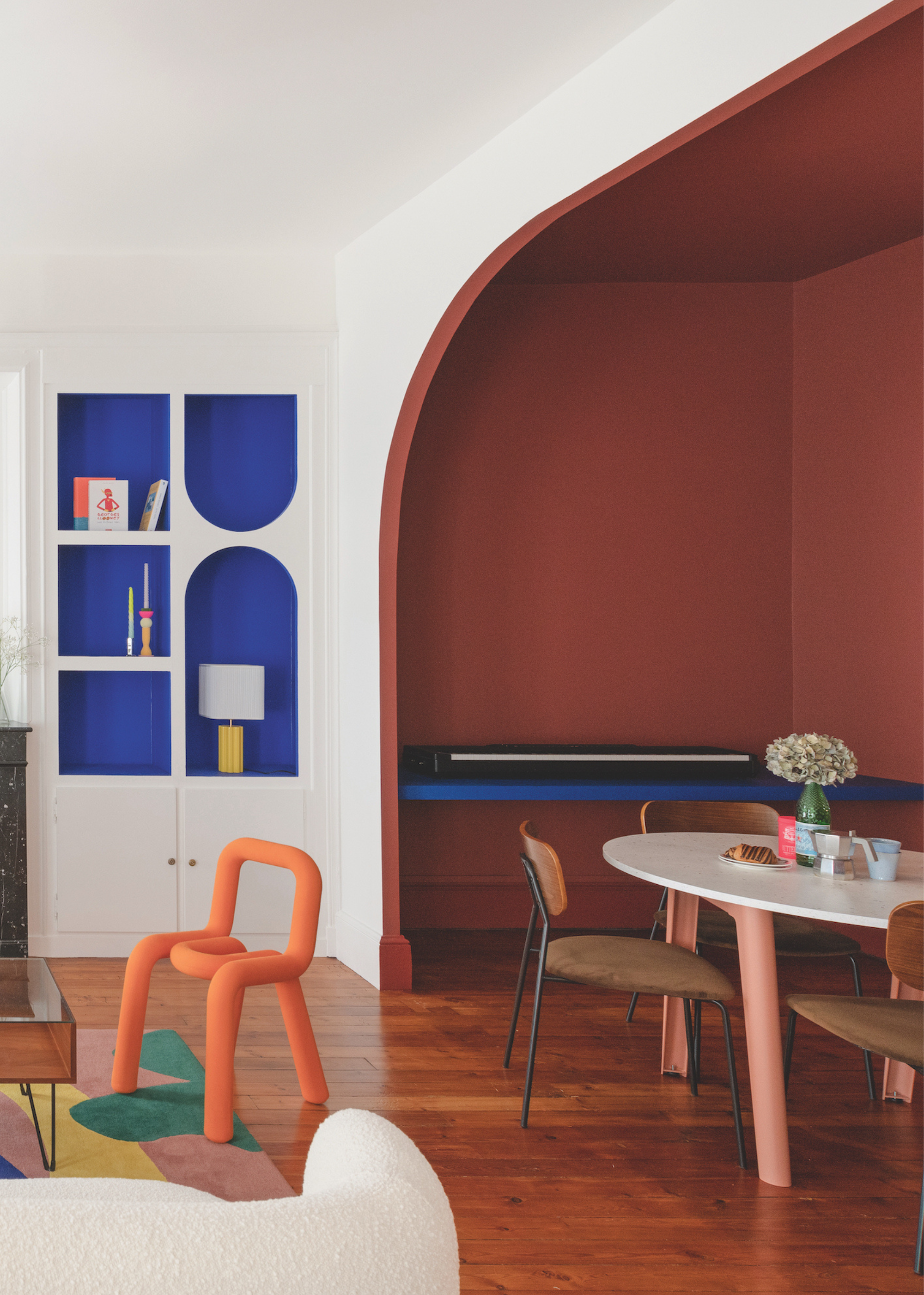 A modern living space with bright colors in a white-painted background and a color drenched area to the right, where the dining area is. There are wooden floors with a bright orange accent chair.