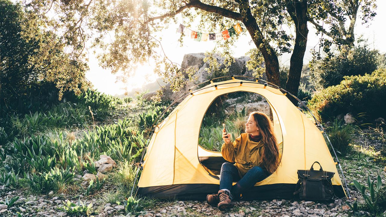 best backpacking tent: Young woman hiker uses mobile phone to check news sitting in small tent in tourist camp in highland against sea and mountains at bright sunset