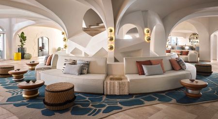 A hotel lounge with vaulted ceilings around which wraps a modular banquette seat in white leather, turquoise blue and gray rugs, wooden, brass, and silver furniture, and rattan puffs.