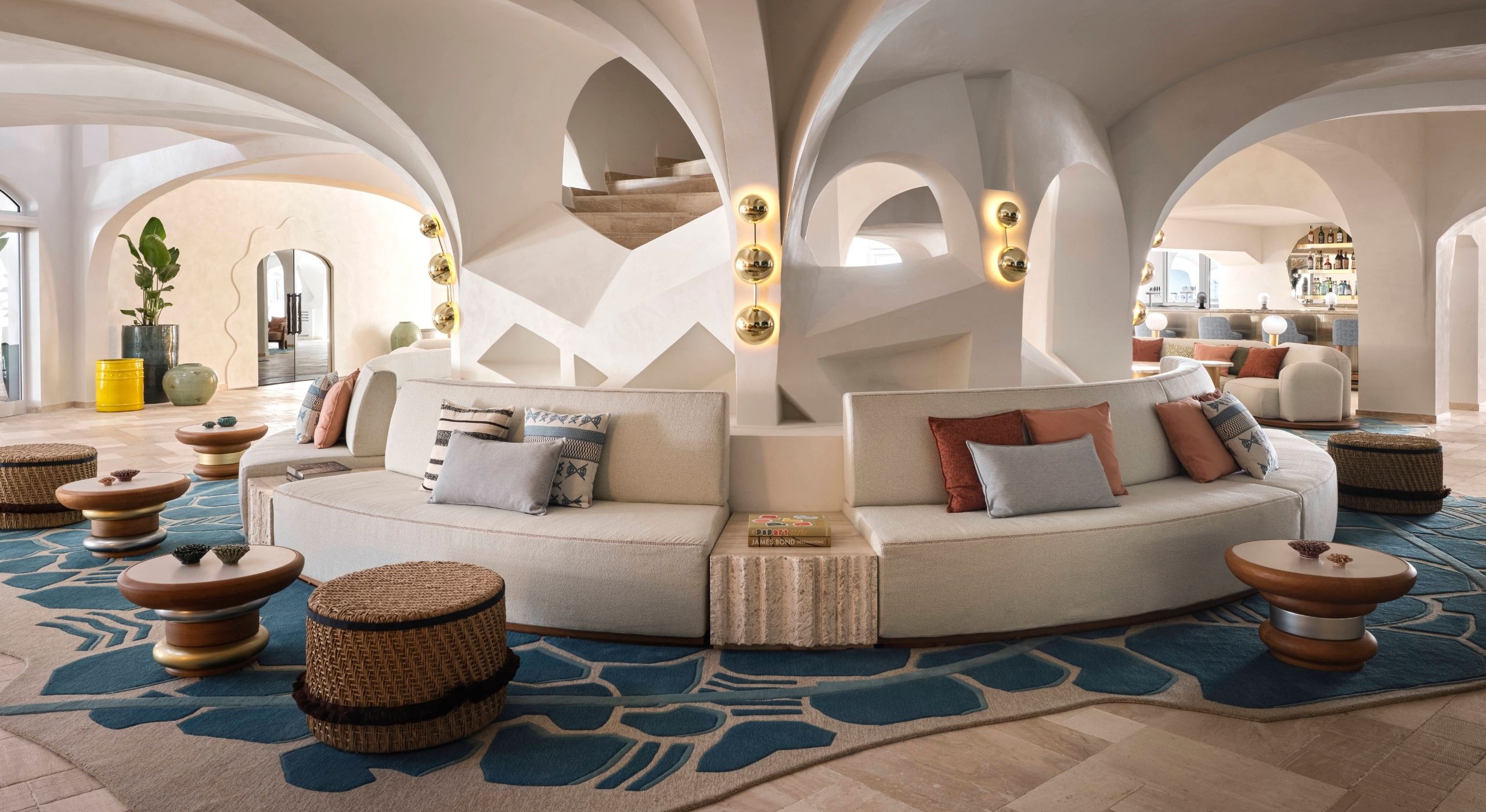 A hotel lounge with vaulted ceilings around which wraps a modular banquette seat in white leather, turquoise blue and gray rugs, wooden, brass, and silver furniture, and rattan puffs.
