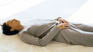 A woman in a gray sweatsuit lies on her back in bed with her hands placed on her pelvis.
