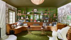 Living room with green painted ceiling and wall, open shelving, wallpaper mural, green rug and large U-shaped brown sofa with modern armchair and two ornate iron side tables
